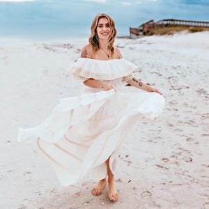 Puede incluir: Vestido blanco con hombros descubiertos, cuello con volantes y falda fluida. El vestido tiene un ribete color crema y se lleva en una playa de arena. El vestido es de estilo bohemio.