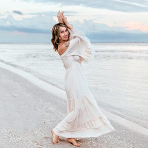 Puede incluir: Una mujer con un vestido largo blanco roto con mangas con volantes y una falda escalonada est&aacute; de pie en una playa de arena. El vestido tiene un estilo bohemio fluido. El fondo presenta el oc&eacute;ano y un cielo nublado.