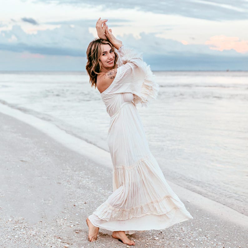 Puede incluir: Una mujer con un vestido largo blanco roto con mangas con volantes y una falda escalonada est&aacute; de pie en una playa de arena. El vestido tiene un estilo bohemio fluido. El fondo presenta el oc&eacute;ano y un cielo nublado.