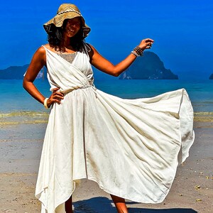 May include: A woman wearing a white maxi dress with a straw hat and a beaded bracelet, standing on a sandy beach with blue water and islands in the background.