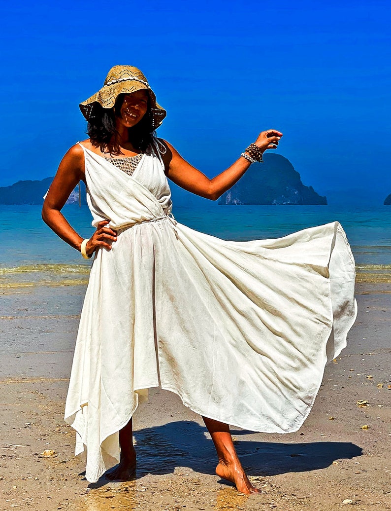 May include: A woman wearing a white maxi dress with a straw hat and a beaded bracelet, standing on a sandy beach with blue water and islands in the background.