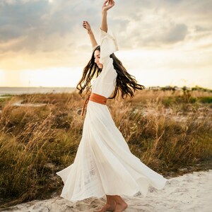 May include: A woman in a white maxi dress with a brown leather belt dances in a field of tall grass. The dress has a flowing skirt and the woman's hair is long and flowing.