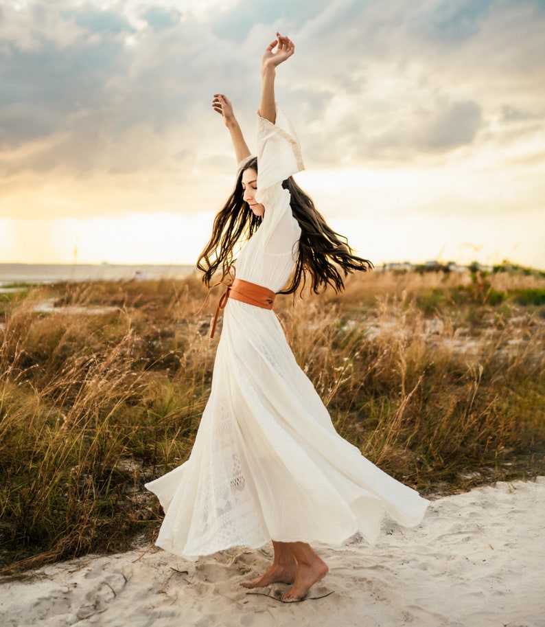 May include: A woman in a white maxi dress with a brown leather belt dances in a field of tall grass. The dress has a flowing skirt and the woman's hair is long and flowing.