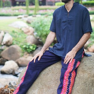 May include: A man wearing a dark blue shirt and dark blue pants with a pink and red patterned trim. He is sitting on a large rock with his legs crossed. The background is a blurred image of green trees and a wooden building.