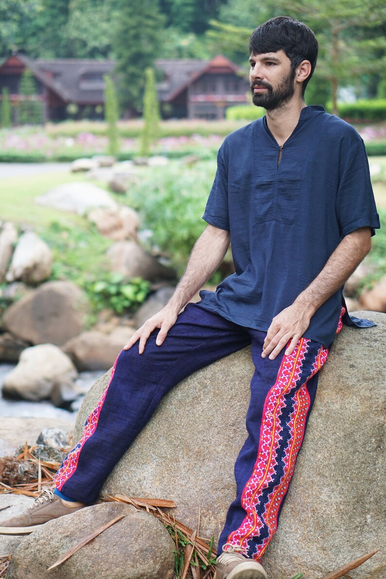 May include: A man wearing a dark blue shirt and dark blue pants with a pink and red patterned trim. He is sitting on a large rock with his legs crossed. The background is a blurred image of green trees and a wooden building.