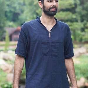 May include: A man wearing a dark blue short-sleeved shirt with a small button at the neckline. He is wearing dark blue pants with a red and pink patterned trim.