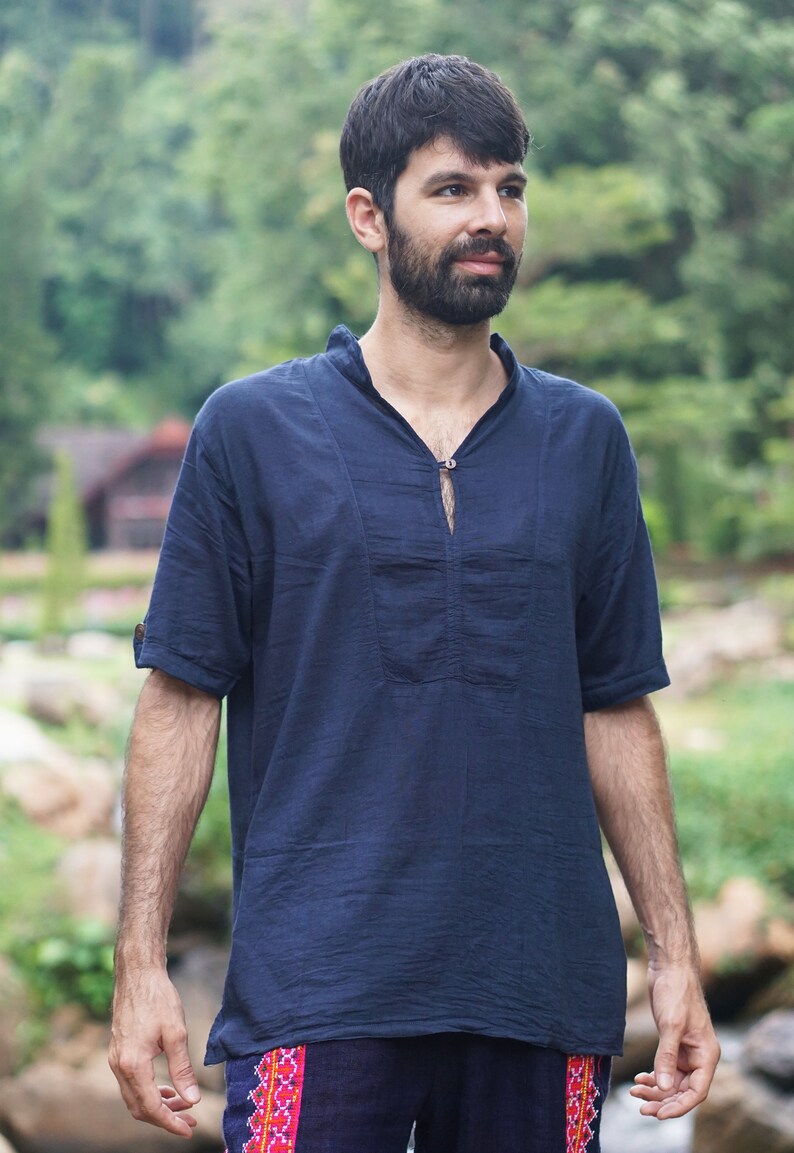 May include: A man wearing a dark blue short-sleeved shirt with a small button at the neckline. He is wearing dark blue pants with a red and pink patterned trim.