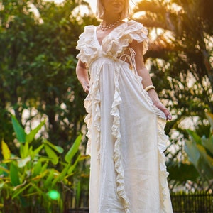 Puede incluir: Un vestido largo color crema con escote en V y volantes. El vestido tiene mangas cortas y una falda fluida. La modelo est&aacute; de pie descalza sobre una superficie de madera, con un fondo de exuberante follaje verde y luz solar.