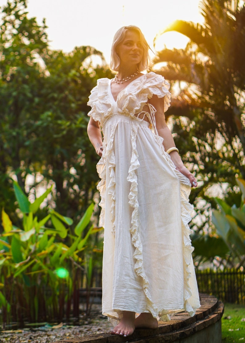 Puede incluir: Un vestido largo color crema con escote en V y volantes. El vestido tiene mangas cortas y una falda fluida. La modelo est&aacute; de pie descalza sobre una superficie de madera, con un fondo de exuberante follaje verde y luz solar.