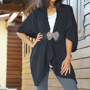 K&ouml;nnte beinhalten: Ein schwarzer Kimono-Cardigan mit gerafftem Saum, getragen mit einem wei&szlig;en Oberteil und grauen Leggings. Ein G&uuml;rtel mit einem Schmuck-Schleifenverschluss wird in der Taille getragen.
