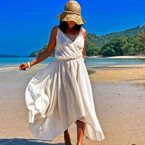 May include: A woman wearing a white maxi dress and a straw hat walks on a sandy beach. The dress has a flowing, asymmetrical hemline. The beach is bordered by lush green trees and a blue sky.