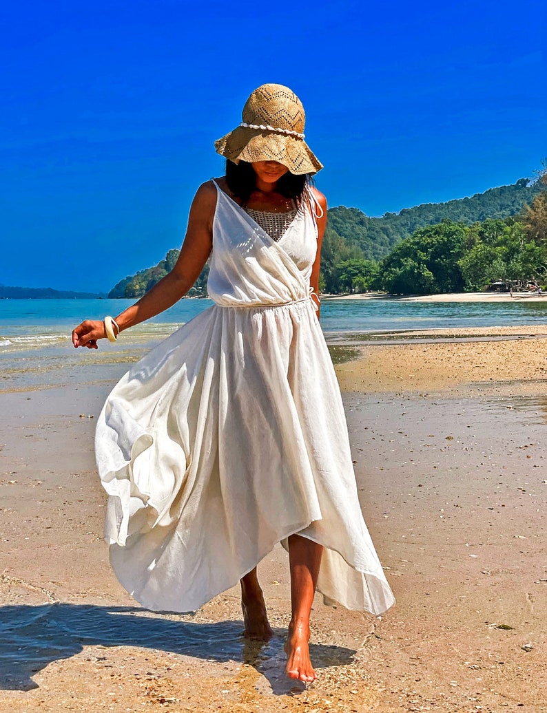 May include: A woman wearing a white maxi dress and a straw hat walks on a sandy beach. The dress has a flowing, asymmetrical hemline. The beach is bordered by lush green trees and a blue sky.