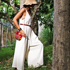 May include: A woman wearing a white jumpsuit with a wide leg, a brown straw hat, and a small, colorful embroidered bag. She is standing in a forest setting.