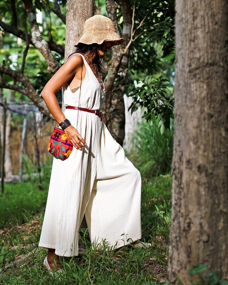 May include: A woman wearing a white jumpsuit with a wide leg, a brown straw hat, and a small, colorful embroidered bag. She is standing in a forest setting.
