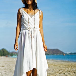 May include: A woman wearing a white dress with a straw hat and a beaded necklace stands on a sandy beach. The ocean is in the background.