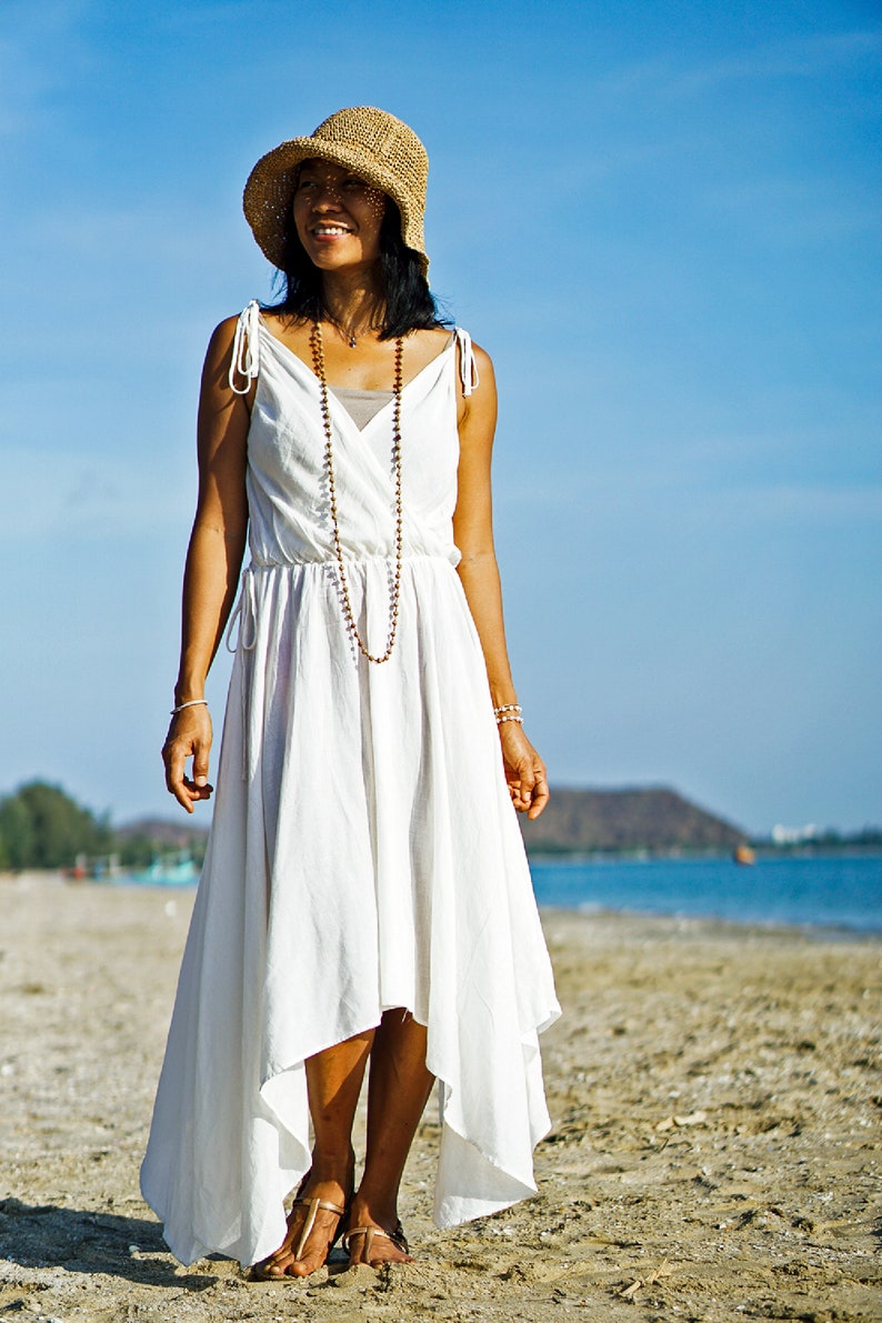 May include: A woman wearing a white dress with a straw hat and a beaded necklace stands on a sandy beach. The ocean is in the background.