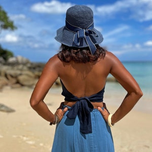 May include: A woman wearing a blue denim skirt and a blue halter top tied in a bow at the back. She is wearing a blue sun hat with a gold ribbon.