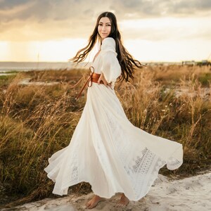 May include: A woman in a white maxi dress with lace detailing and a brown leather belt poses in a field of tall grass. The dress is flowing in the wind, and the woman is smiling at the camera.