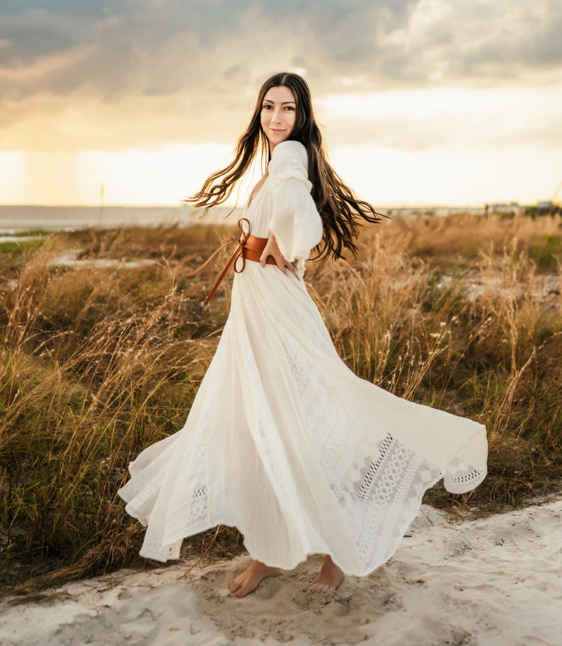 May include: A woman in a white maxi dress with lace detailing and a brown leather belt poses in a field of tall grass. The dress is flowing in the wind, and the woman is smiling at the camera.