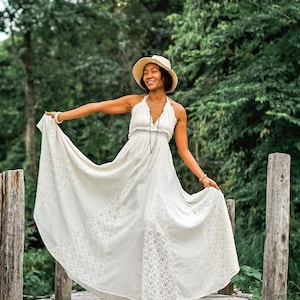 May include: A cream-colored halter maxi dress with lace detailing. The dress has a flowing skirt and is being held up to show the design. A straw hat and jewelry complete the look. The background is a wooden bridge and lush greenery.