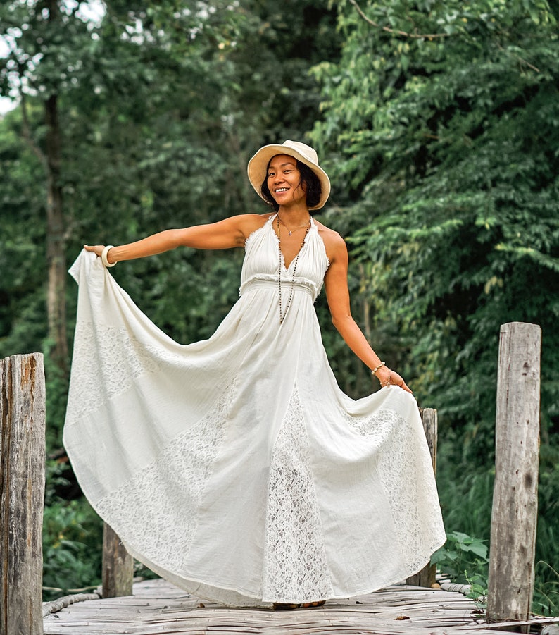 May include: A cream-colored halter maxi dress with lace detailing. The dress has a flowing skirt and is being held up to show the design. A straw hat and jewelry complete the look. The background is a wooden bridge and lush greenery.