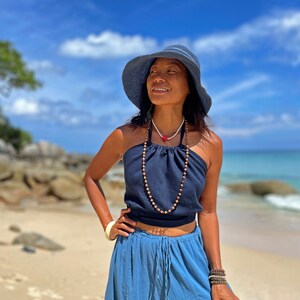 May include: A woman wearing a blue halter top with a wooden bead necklace, a blue skirt, and a blue sun hat. She is standing on a beach with a blue sky and white clouds in the background.