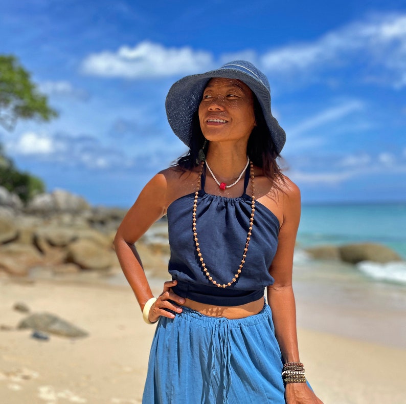 May include: A woman wearing a blue halter top with a wooden bead necklace, a blue skirt, and a blue sun hat. She is standing on a beach with a blue sky and white clouds in the background.