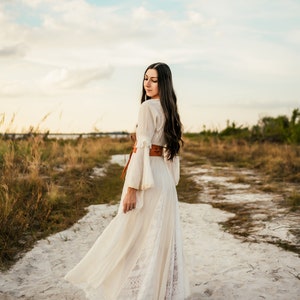 May include: A woman in a white maxi dress with lace detailing and a brown leather belt walks on a sandy path. The dress has a flowing skirt and billowing sleeves. The path is surrounded by tall grass.