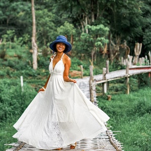 May include: A woman wearing a white maxi dress with a lace panel and a blue hat stands on a wooden bridge. The dress has a halter neckline and flows gracefully. The background features lush green foliage and a wooden bridge.