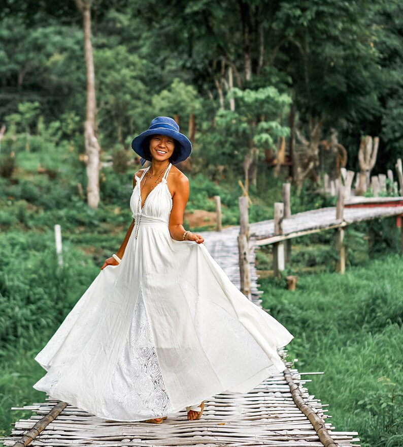 May include: A woman wearing a white maxi dress with a lace panel and a blue hat stands on a wooden bridge. The dress has a halter neckline and flows gracefully. The background features lush green foliage and a wooden bridge.