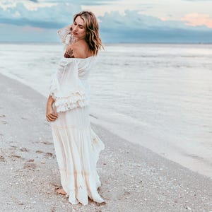 Puede incluir: Un vestido crema con hombros descubiertos, mangas largas con volantes y falda fluida. El vestido se lleva en una playa con el oc&eacute;ano y el cielo de fondo. El vestido tiene un estilo bohemio.