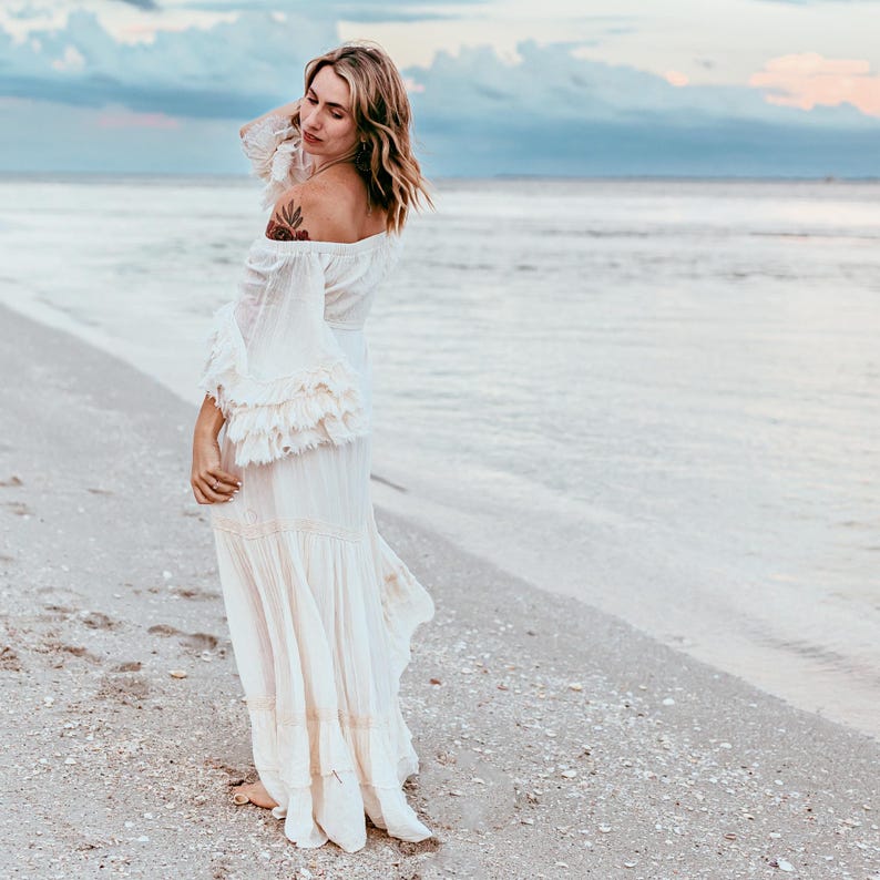 Puede incluir: Un vestido crema con hombros descubiertos, mangas largas con volantes y falda fluida. El vestido se lleva en una playa con el oc&eacute;ano y el cielo de fondo. El vestido tiene un estilo bohemio.