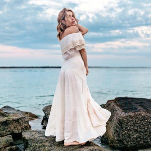 Puede incluir: Una mujer con un vestido blanco con hombros descubiertos y falda escalonada est&aacute; de pie sobre rocas cerca del oc&eacute;ano. El vestido tiene detalles de encaje en el escote. El cielo es azul con nubes.