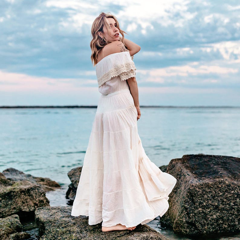 Puede incluir: Una mujer con un vestido blanco con hombros descubiertos y falda escalonada est&aacute; de pie sobre rocas cerca del oc&eacute;ano. El vestido tiene detalles de encaje en el escote. El cielo es azul con nubes.