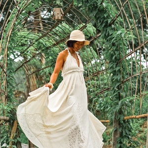 May include: A woman wearing a cream-colored halter maxi dress with lace trim, a beige sun hat, and sandals, walking on a wooden bridge. The dress flows in the wind. The background features a green archway covered in vines.