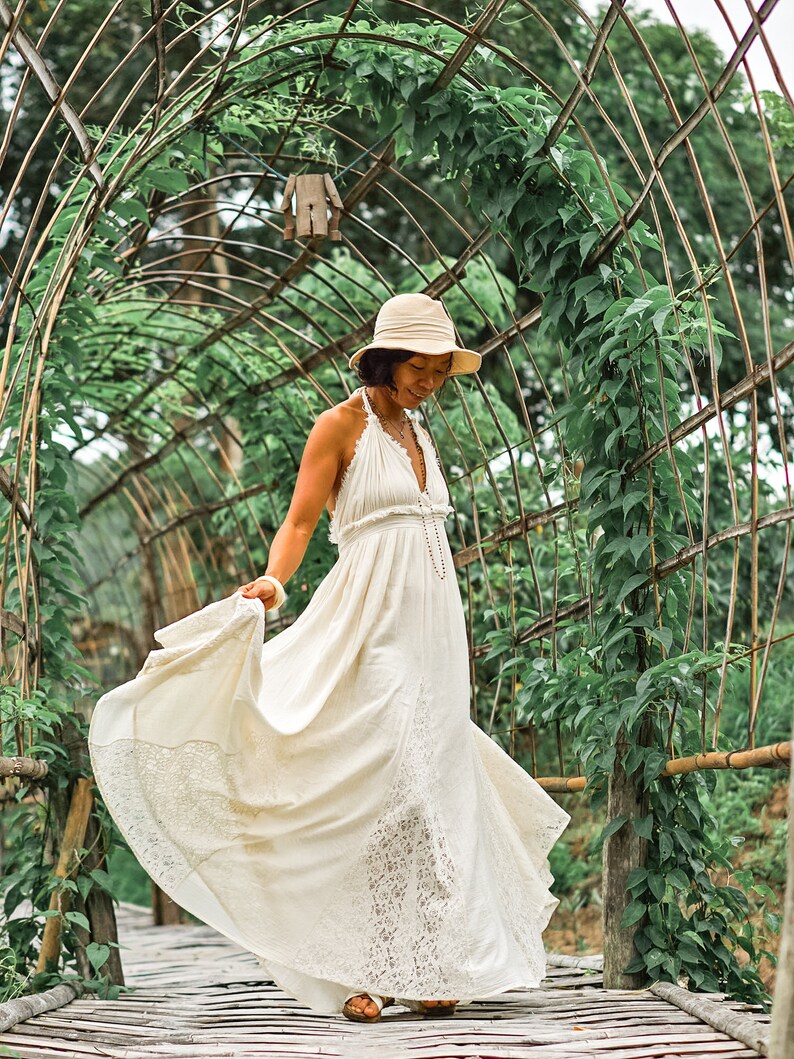 May include: A woman wearing a cream-colored halter maxi dress with lace trim, a beige sun hat, and sandals, walking on a wooden bridge. The dress flows in the wind. The background features a green archway covered in vines.