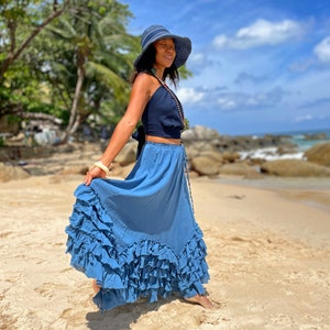 May include: A woman wearing a blue and white hat, a dark blue halter top, and a long, flowing blue skirt with multiple ruffles. She is standing on a sandy beach with rocks and the ocean in the background.