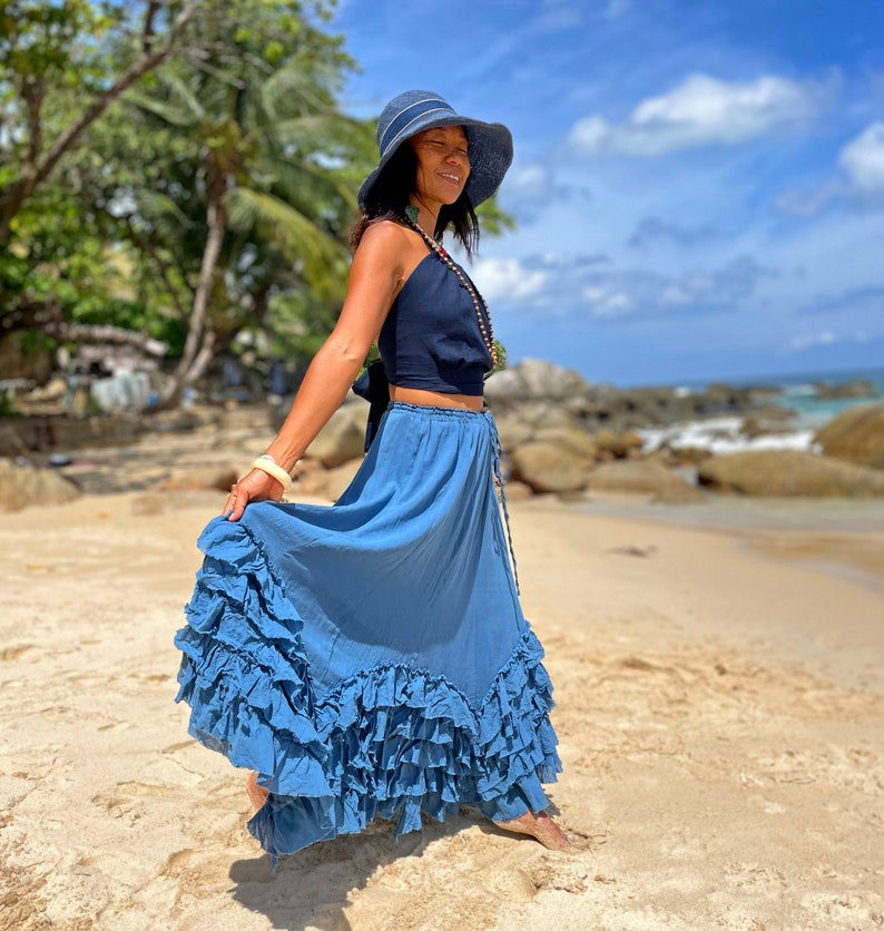 May include: A woman wearing a blue and white hat, a dark blue halter top, and a long, flowing blue skirt with multiple ruffles. She is standing on a sandy beach with rocks and the ocean in the background.