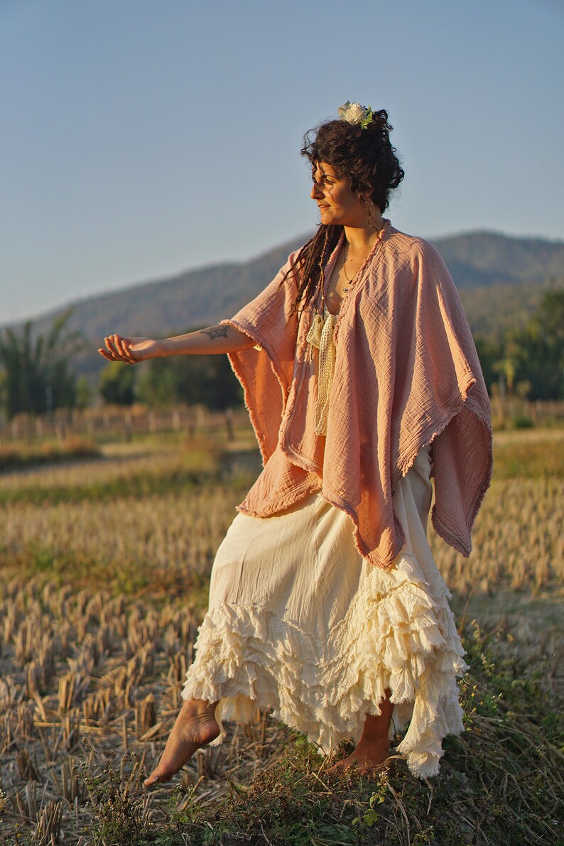 May include: A woman wearing a pink and white flowy dress with a white flower in her hair, standing in a field of dry grass.