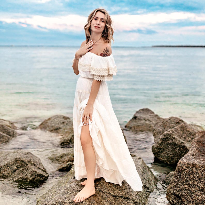 Puede incluir: Un vestido largo blanco con hombros descubiertos y ribete de encaje, de pie sobre rocas cerca del oc&eacute;ano. El vestido tiene una falda fluida y una abertura alta. El cielo es azul con nubes.