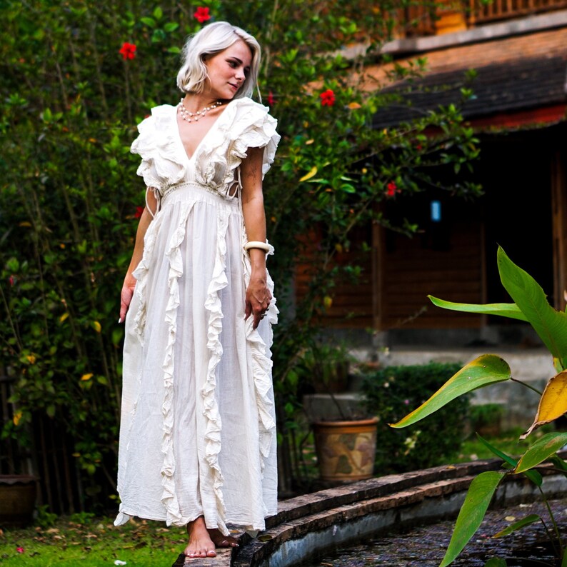 Puede incluir: Un vestido largo color crema con volantes. El vestido presenta un escote en V profundo y una falda fluida. La modelo est&aacute; de pie en una pasarela de ladrillos en un jard&iacute;n. El vestido es de estilo bohemio.