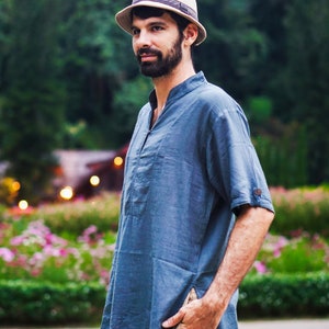 May include: A man wearing a blue short-sleeved shirt with a light brown hat. He is standing in front of a green background with flowers.