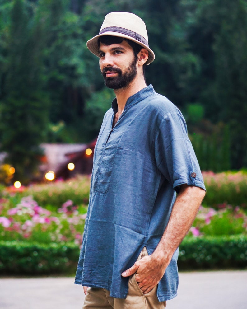 May include: A man wearing a blue short-sleeved shirt with a light brown hat. He is standing in front of a green background with flowers.