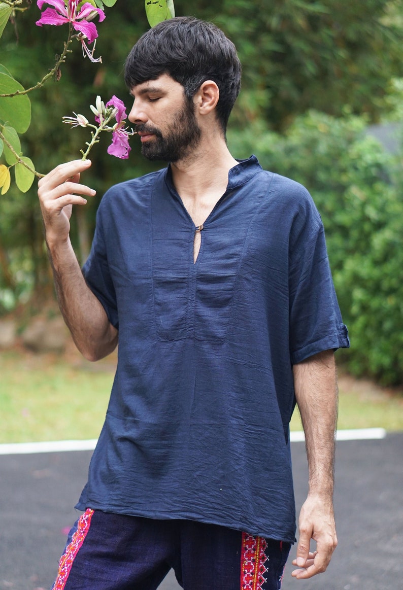 May include: A man wearing a dark blue short-sleeved shirt with a mandarin collar and a single button closure. He is holding a pink flower in his hand and smelling it.