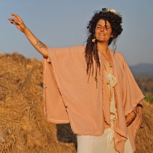 May include: A woman with long brown dreadlocks wears a light pink kimono with a white crochet top underneath. She is smiling and has a flower crown in her hair.