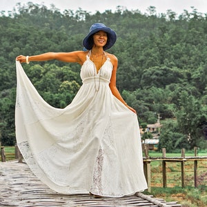 May include: A woman wearing a white halter dress and a blue hat stands on a wooden bridge. The dress has a flowing skirt with lace detailing. The background features lush green trees and a cloudy sky.