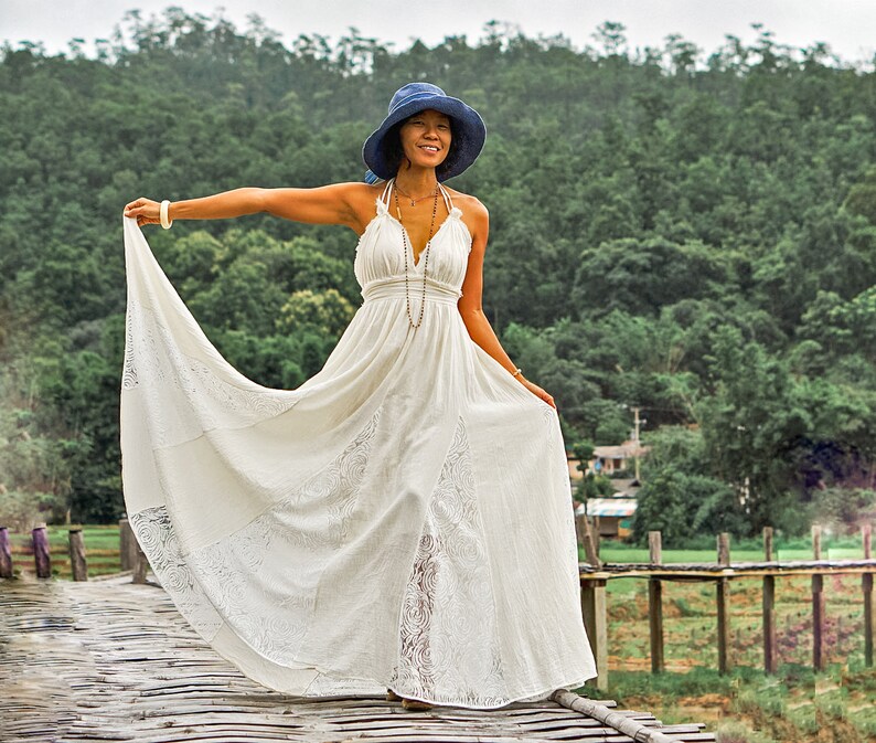 May include: A woman wearing a white halter dress and a blue hat stands on a wooden bridge. The dress has a flowing skirt with lace detailing. The background features lush green trees and a cloudy sky.