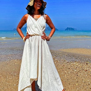 May include: A woman wearing a white linen dress and a straw hat stands on a sandy beach. The dress has a wrap style and a flowing skirt. The woman is smiling and looking up at the sky.