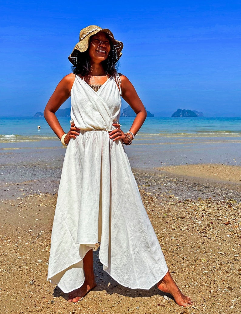 May include: A woman wearing a white linen dress and a straw hat stands on a sandy beach. The dress has a wrap style and a flowing skirt. The woman is smiling and looking up at the sky.