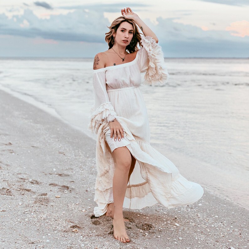 Puede incluir: Vestido color crema con hombros descubiertos, mangas con volantes y falda fluida. El vestido se lleva en una playa de arena con el oc&eacute;ano al fondo. El vestido tiene cintura entallada y falda escalonada.